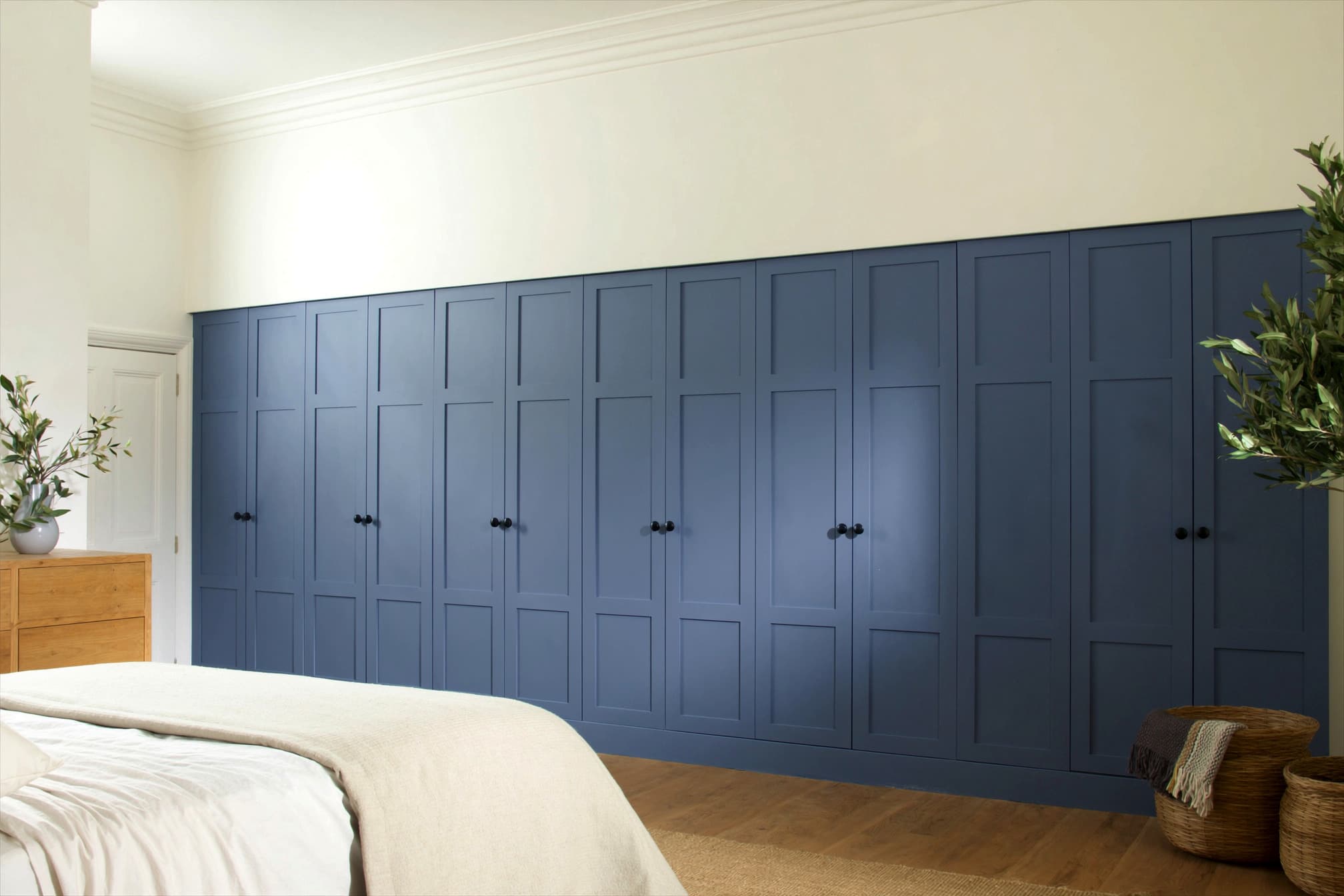 A fitted bedroom with matt denim-blue shaker wardrobes and black knobs, on a warm oak floor