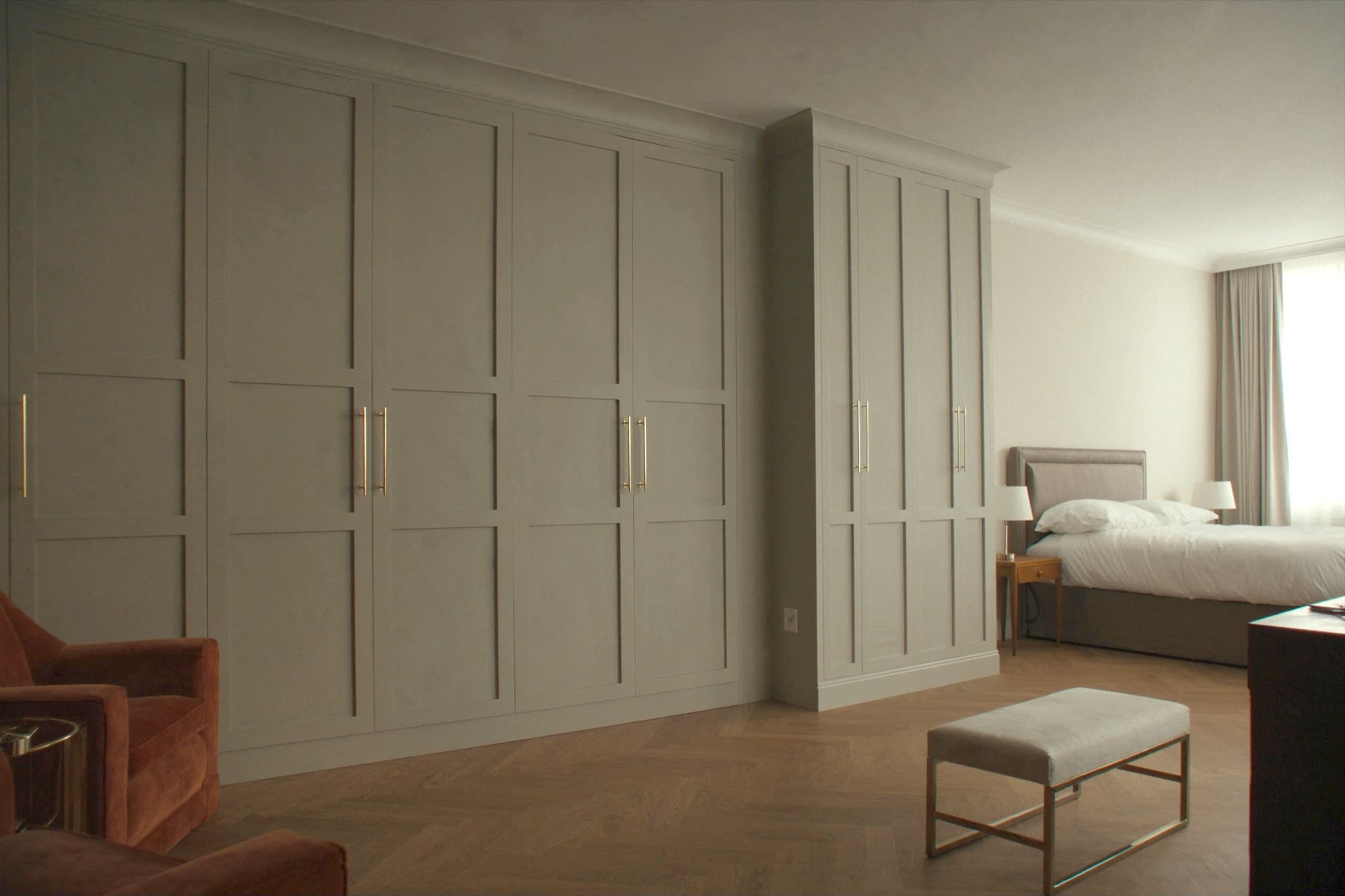 A fitted bedroom with a corner run of sage-grey shaker wardrobes with a moulded cornice and brass bar handles