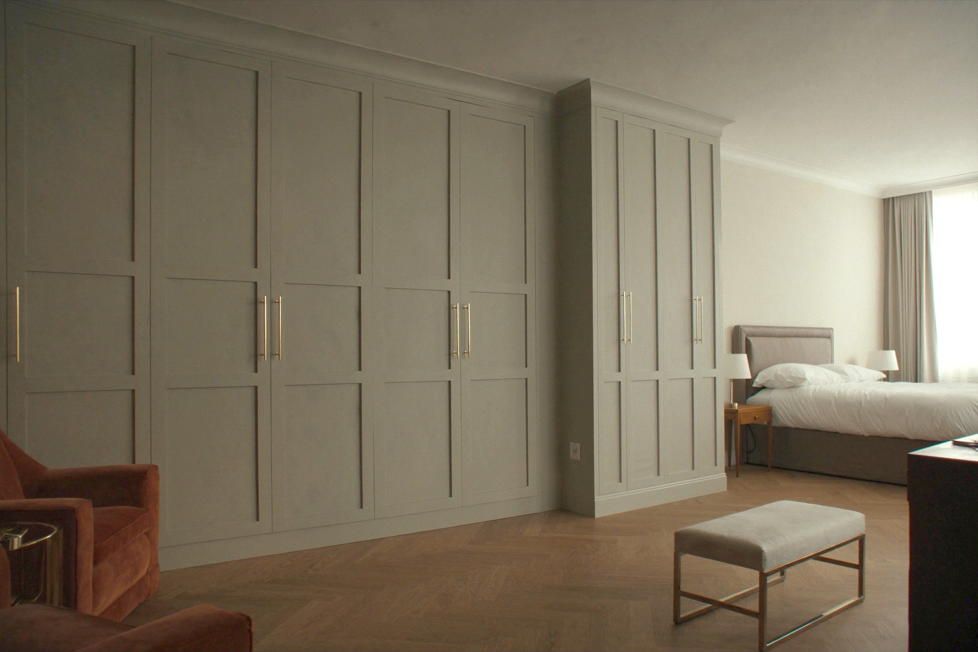 A fitted bedroom with a corner run of sage-grey shaker wardrobes with a moulded cornice and brass bar handles, on a herringbone oak floor