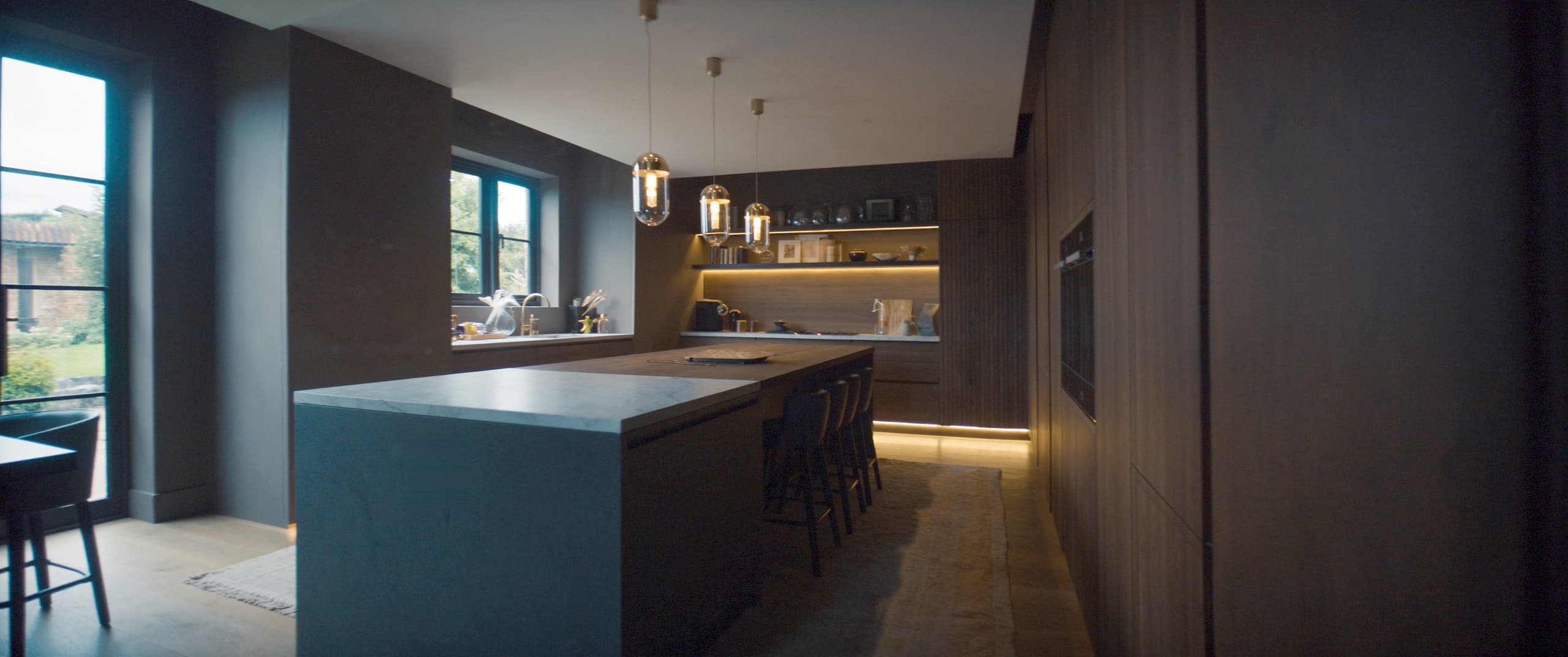 A bespoke charcoal and walnut kitchen at dusk, the island lit by aged brass pendants