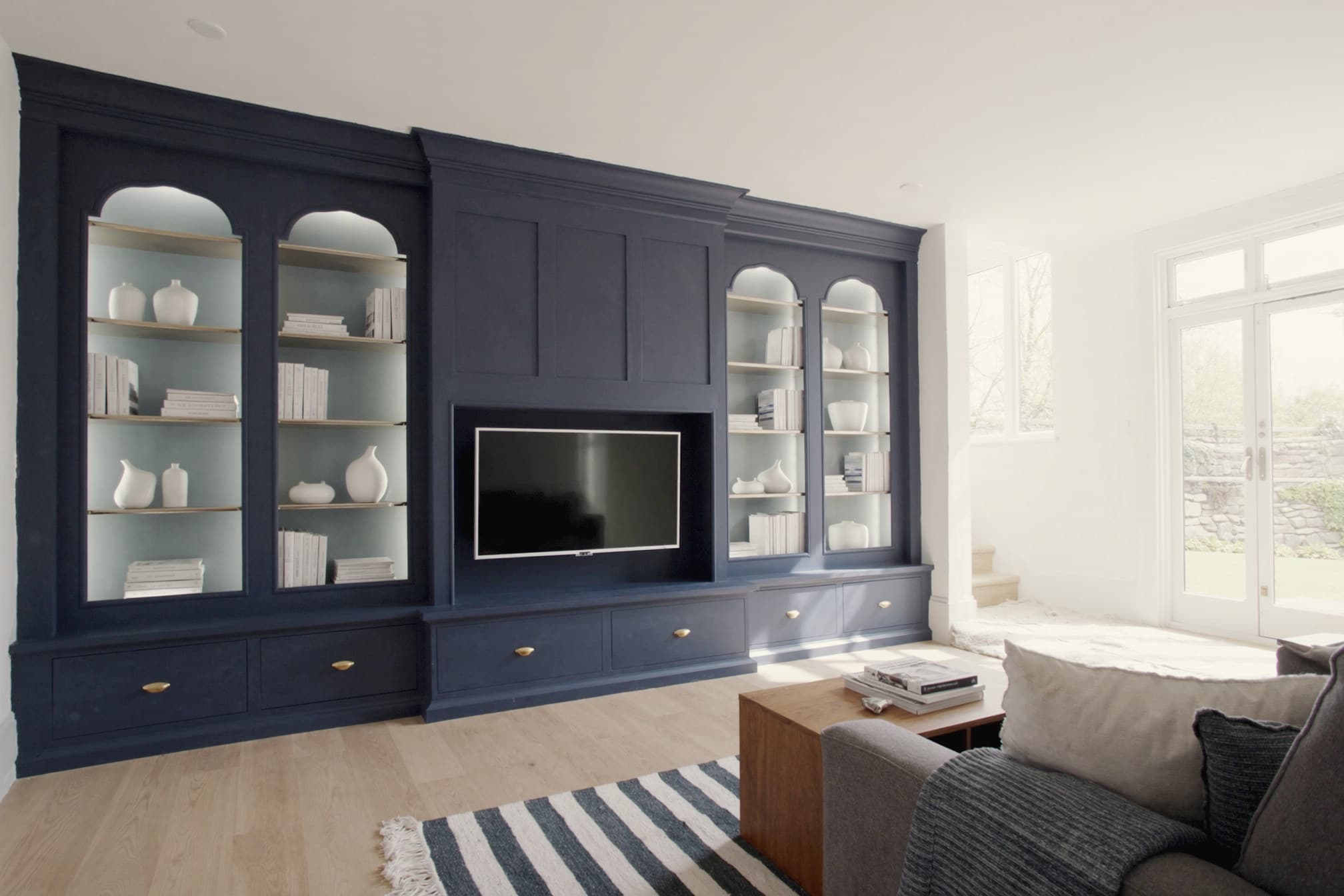 A navy shaker media wall filling a living-room wall, with arched glazed display bays, open shelving and base cupboards with brass knobs