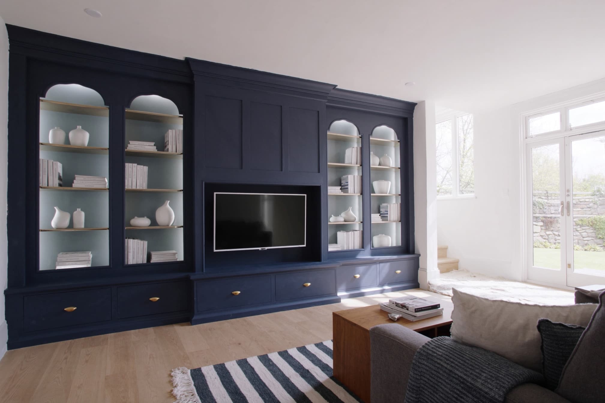 A navy shaker media wall filling a living-room wall, with arched glazed display bays, open shelving and base cupboards with brass knobs