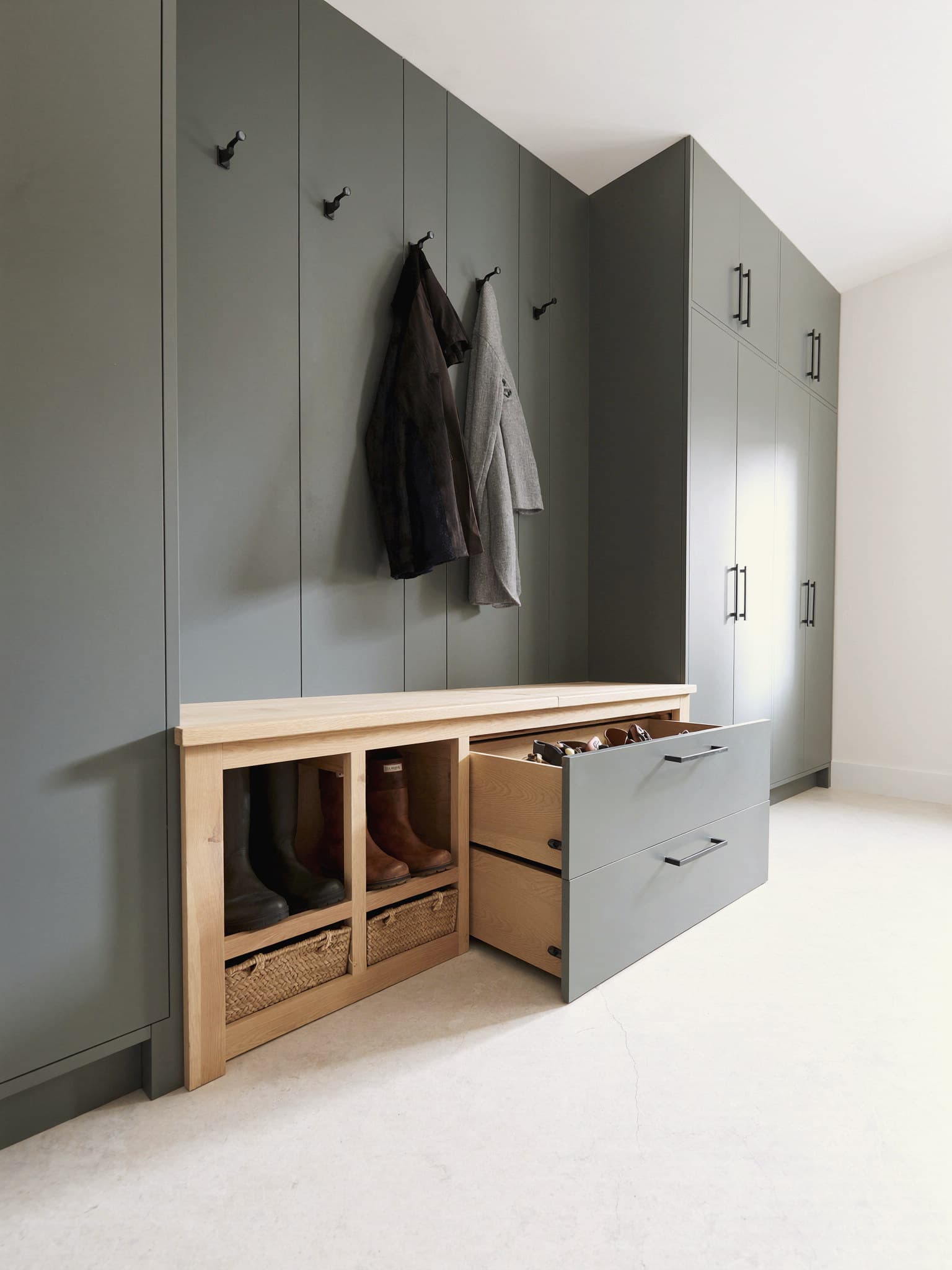 A fitted boot room in grey-green: an oak bench seat, black coat hooks, open cubbies holding boots and baskets, and deep oak-lined drawers