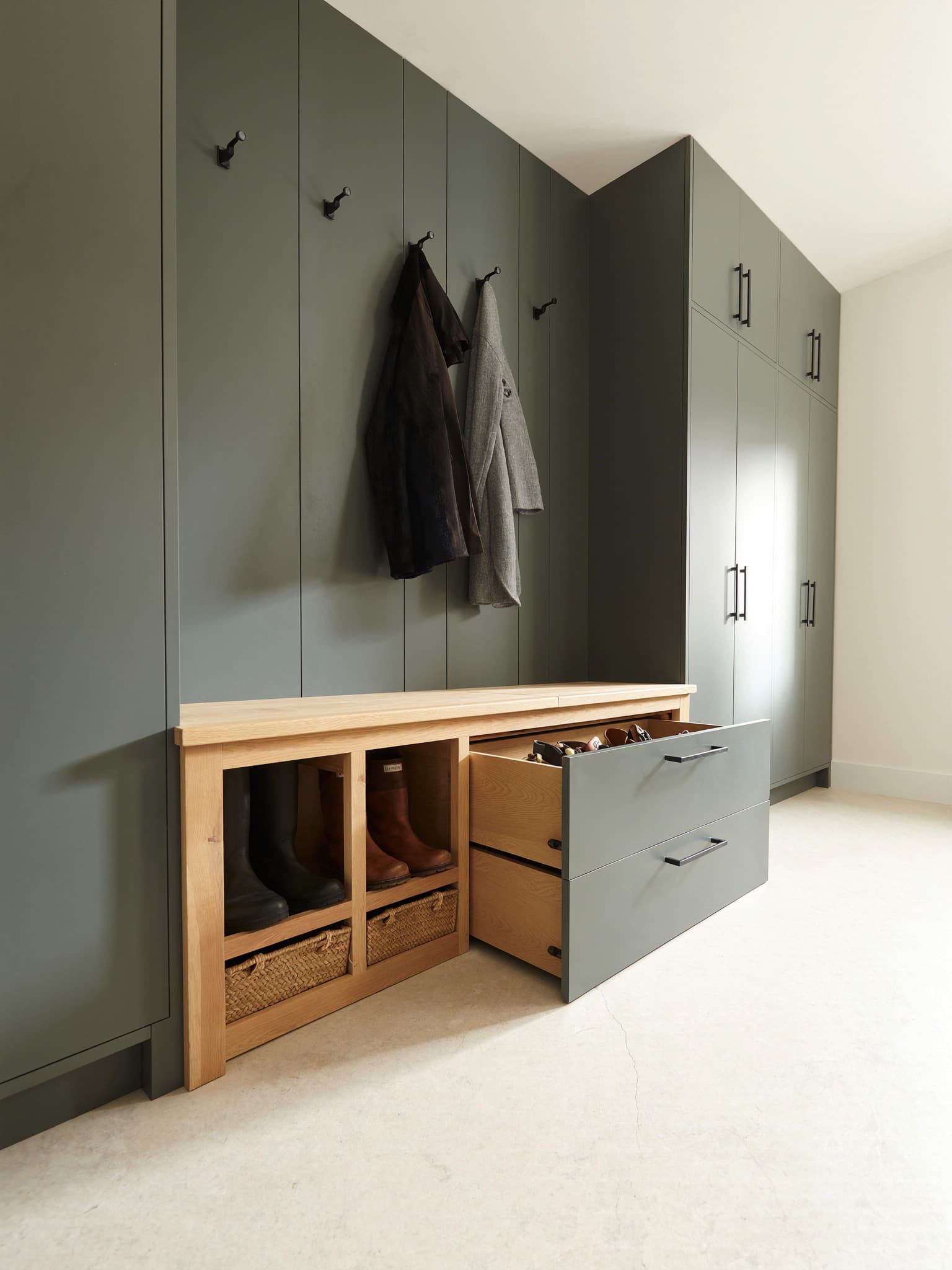 A fitted boot room in grey-green: an oak bench seat, black coat hooks, open cubbies holding boots and baskets, and deep oak-lined drawers