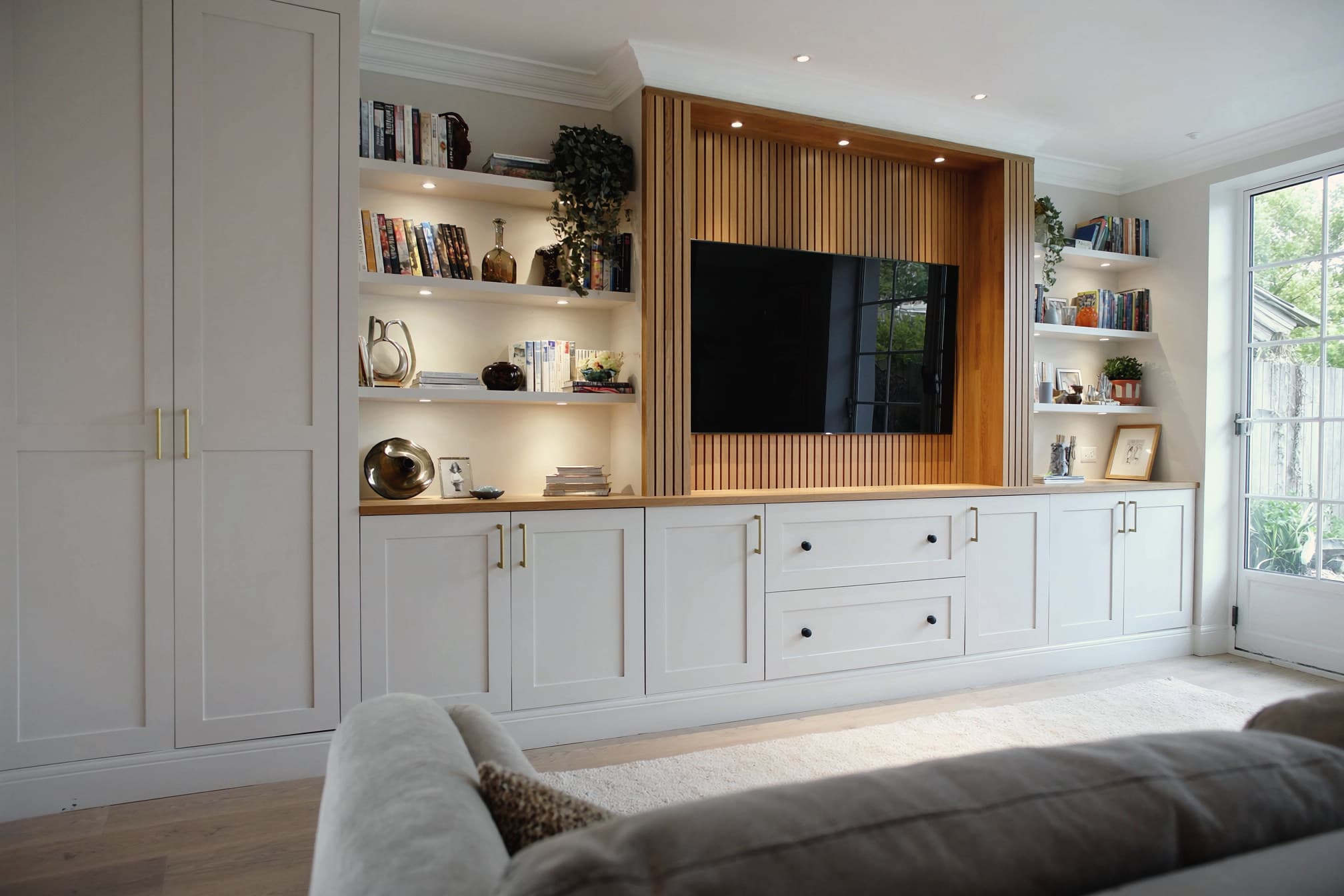 A media wall in warm greige with an oak slat television recess and lit open shelving