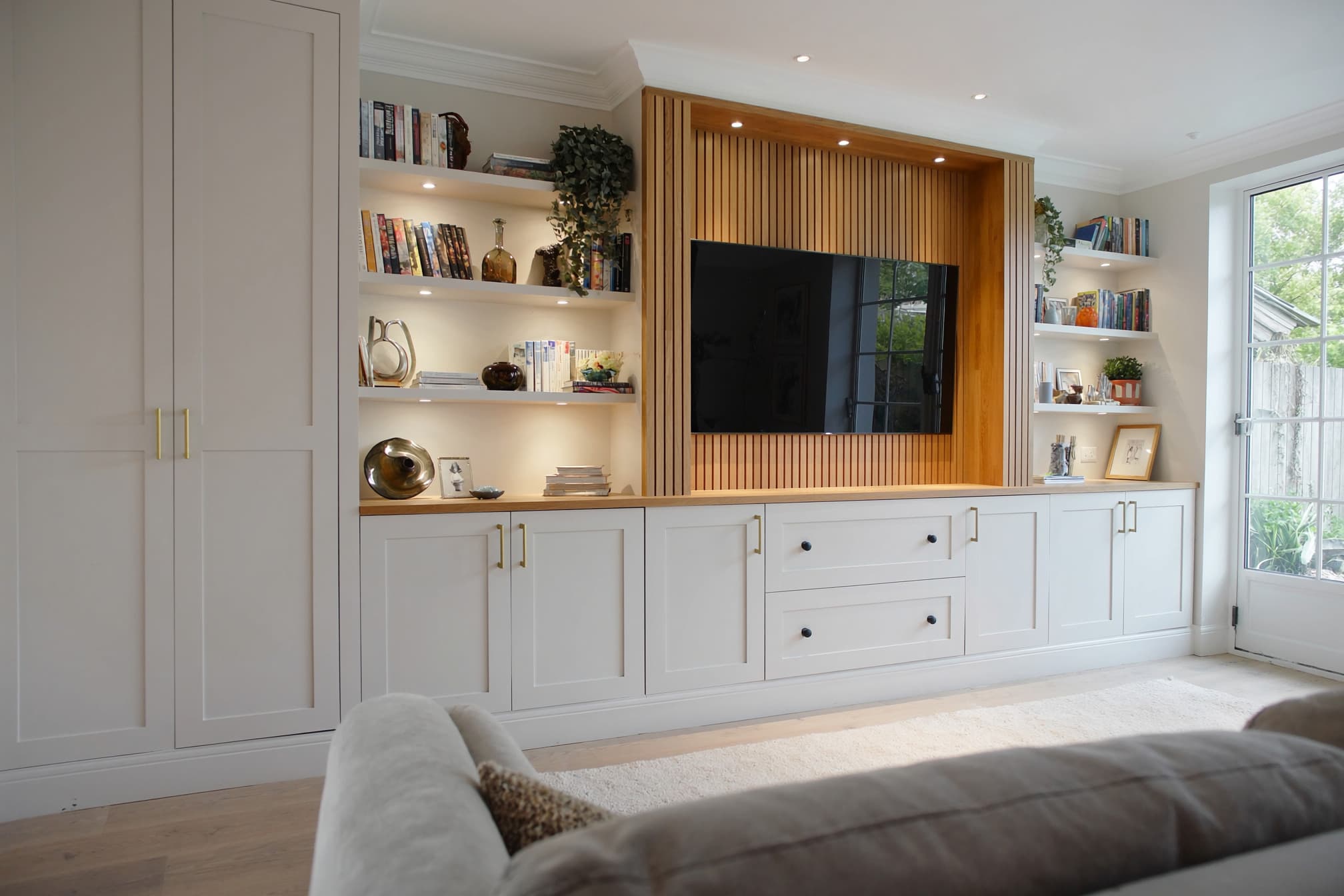 A media wall in warm greige with an oak slat television recess and lit open shelving