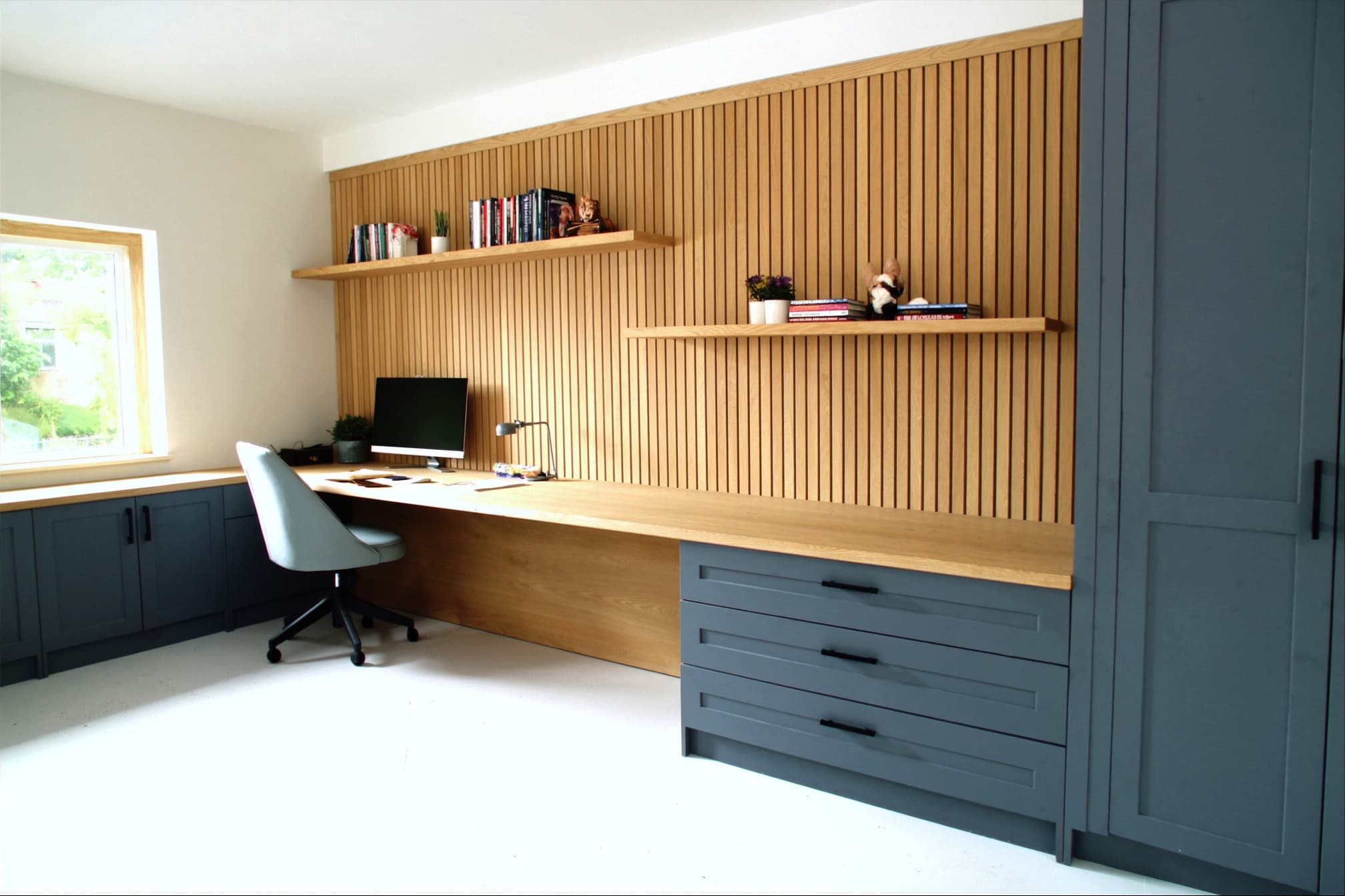 A fitted study in pale blue-grey shaker: a long oak desk against an oak slat wall with floating shelves, and a bank of drawers beneath