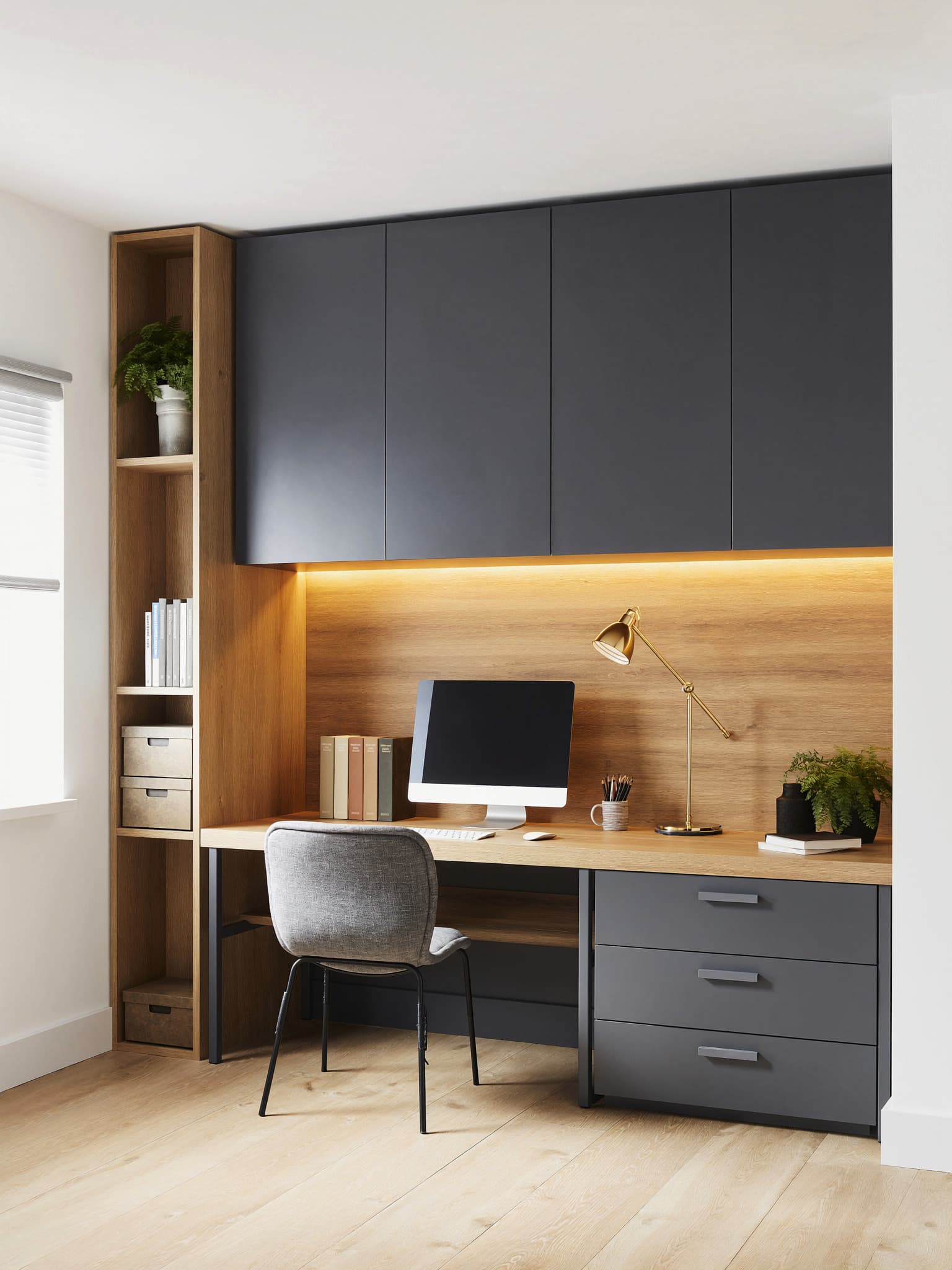A fitted desk bay in graphite grey with a warm oak-lined back panel lit by a concealed LED strip, an oak worktop and a tall oak shelving column