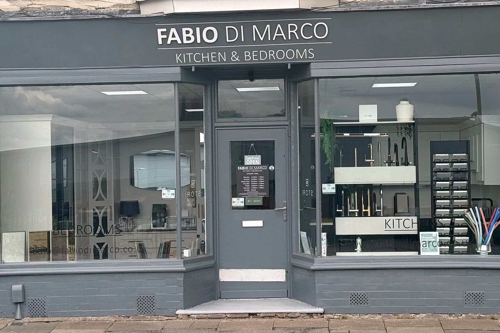 The Fabio Di Marco shop on New Buildings in Hinckley: a grey painted frontage with the name and KITCHEN & BEDROOMS across the fascia, an OPEN sign hanging in the glazed door, fitted bedroom furniture behind the left window and kitchen displays with tap and worktop samples behind the right