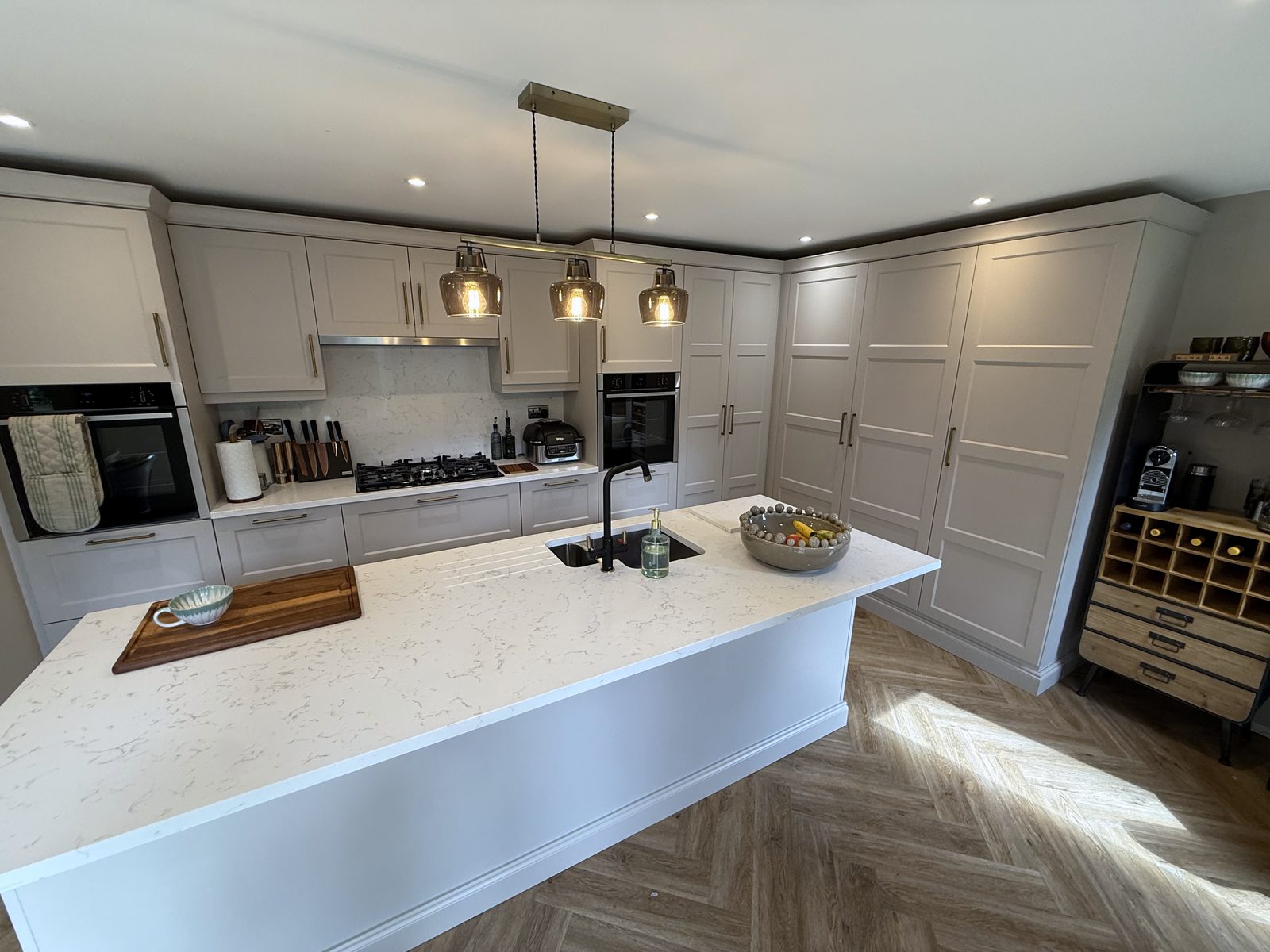 A fitted kitchen by Fabio Di Marco: pale grey shaker doors with brass bar handles, a central island with a white marbled quartz top and a black tap, three smoked-glass pendants over it and a bank of tall larder units, on a herringbone floor