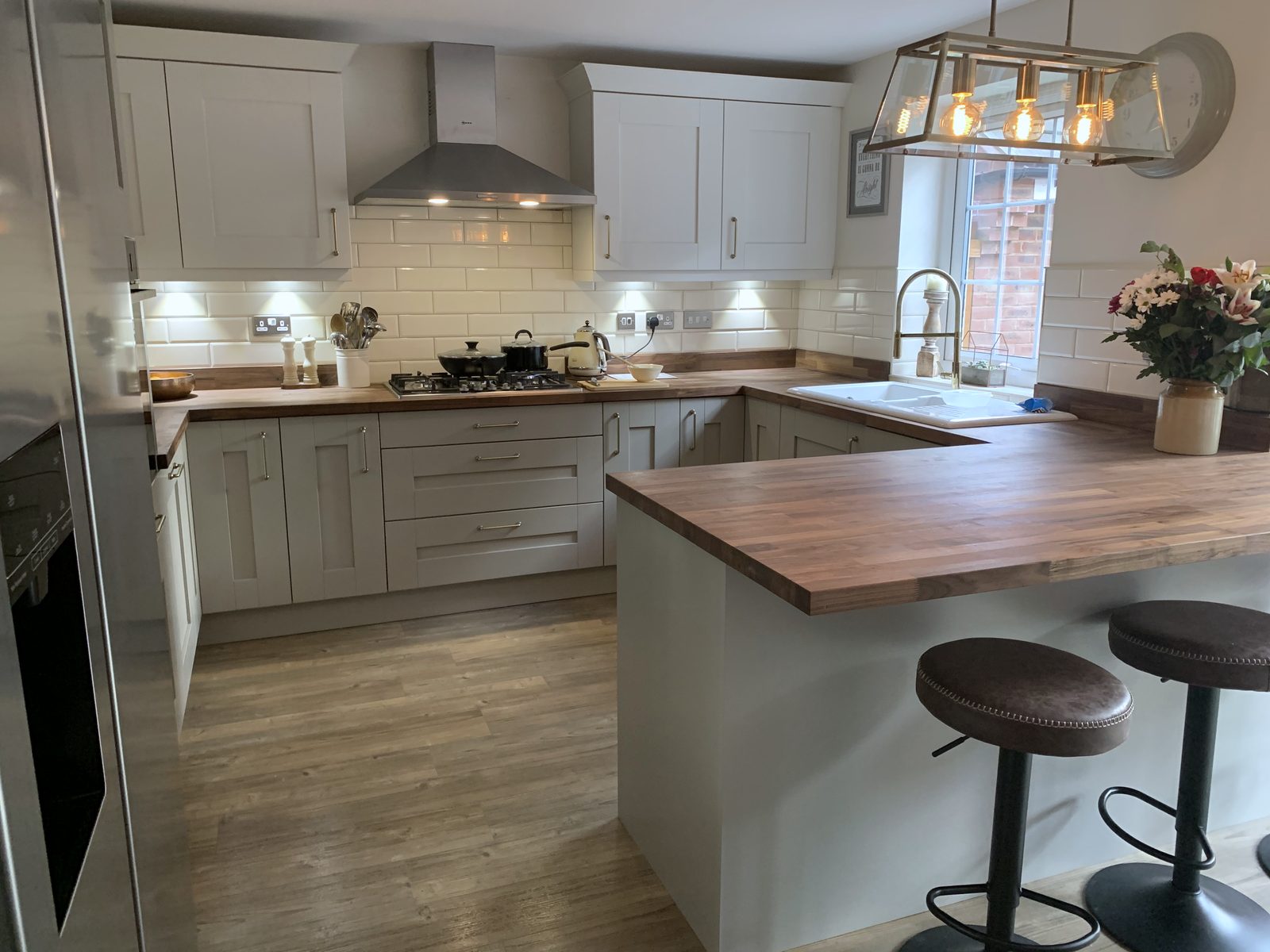 A fitted kitchen by Fabio Di Marco in daylight: pale grey shaker units with a walnut worktop and breakfast bar, cream metro tiles, a brass tap at the window, flowers on the side and two leather stools, on an oak floor